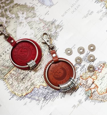 Red and brown leather Vagabond Life keychains with engraved travel rings on a vintage world map.