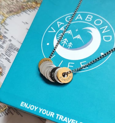 Vagabond Life engraved continent rings on a silver chain necklace over a blue travel box and world map.