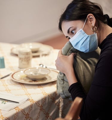 A woman wearing a mask holds a vibrating cushion filled with chai spices at a kitchen table 
