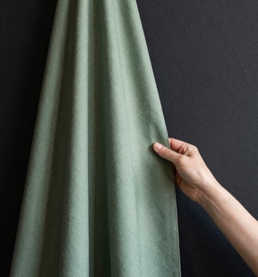 Minimalist close-up of a designer's hand holding a sage green textile swatch against a charcoal black wall, soft natural light.