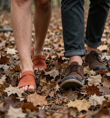 A close-up shot of a couple's feet walking through dry leaves in a North American forest. Earthy terracotta, charcoal, and soft sand colors in a cinematic style.