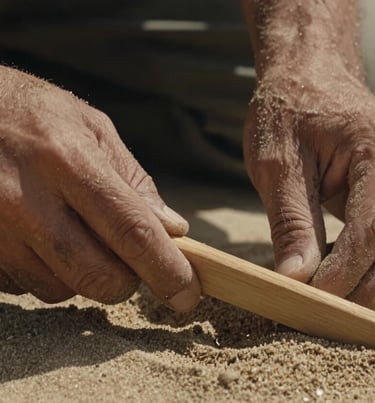 A close up of the artist's hands covered in fine soft sand, holding a simple wooden tool, warm cinematic light.