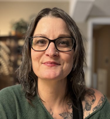 Headshot of Lori Glazebrook smiling  with glasses and shoulder tattoos wearing a green sweater.