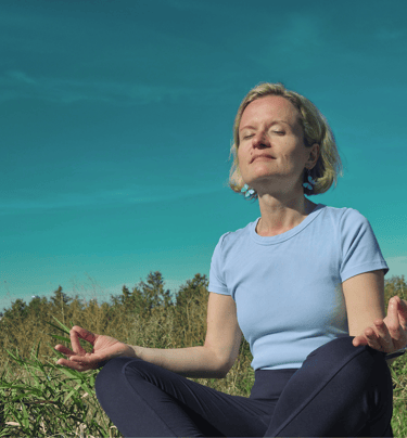 Laura in a meditation lotus position in nature, surrounded by green grass and blue skies.