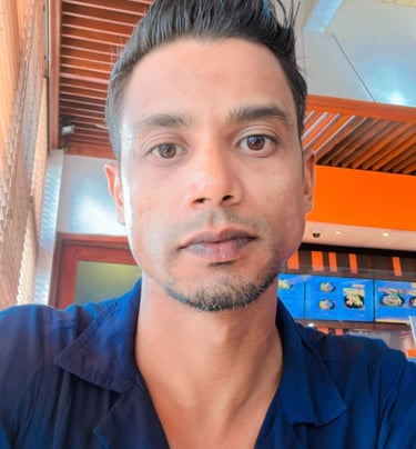 A man with styled dark hair and a blue shirt sitting in a modern cafe with wooden decor.