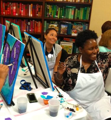 a smiling and laughing group of people sitting at a table painting on canvases