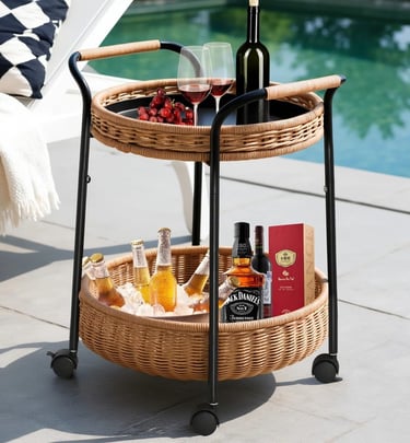 Two-tier wicker outdoor bar cart with wine and bottled drinks on a patio by a luxury swimming pool.