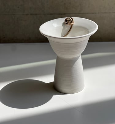 A wheel thrown, white porcelain bowl with an elegant pedestal base. On its rim is a cast bronze knot