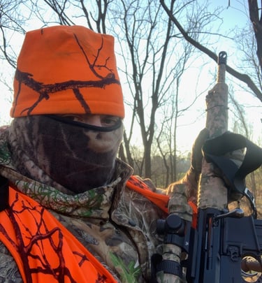 A hunter wearing a blaze orange safety vest and hat while holding a camouflaged rifle in a winter forest.