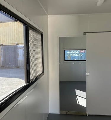 Interior of a white portable site office with a sliding door and windows overlooking a shipping container yard.