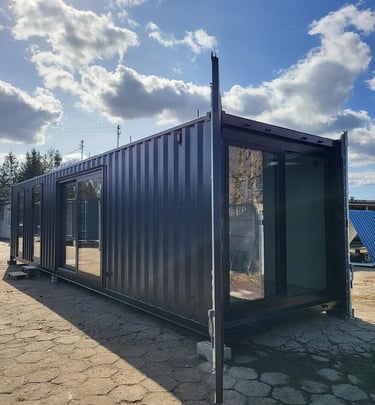 Modern dark grey shipping container home with large glass sliding doors under a bright blue sky.