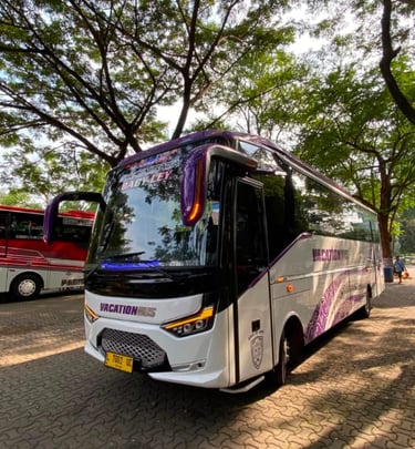 A modern white and purple vacation tour bus parked under leafy green trees on a paved lot.