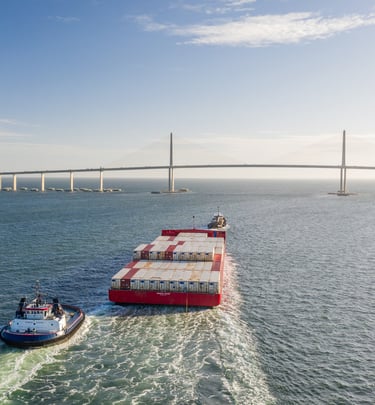 Aerial view of a red cargo ship and tugboat passing under the Sunshine Skyway Bridge in Florida.
