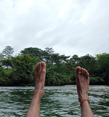 Bare feet relaxing over a flowing river with a background of lush tropical green trees and a cloudy sky.