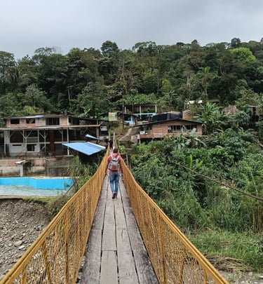 A hiker crosses a narrow wooden suspension bridge towards a hillside village in the tropical rainforest.