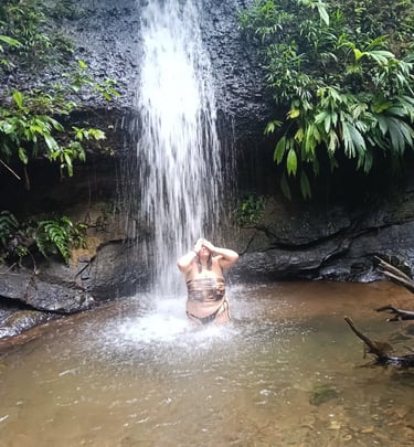 A woman bathing under a natural tropical waterfall surrounded by lush green jungle foliage.