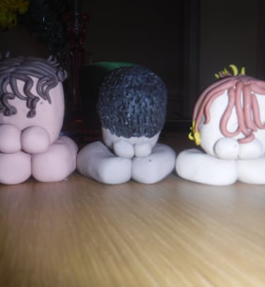 Three small clay figures with different hair textures sit on a wooden table in a dimly lit room.