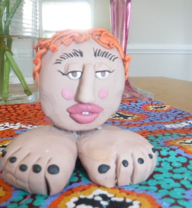 Handmade clay head sculpture with orange hair and feet base on a colorful patterned tablecloth.