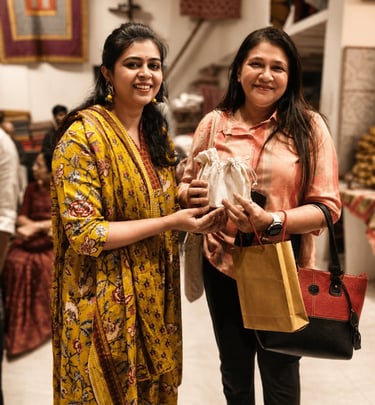 Two women smiling while holding a small fabric gift bag at a traditional Indian clothing boutique.