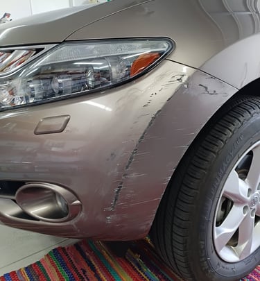 Close-up of a scratched silver car bumper and fender before Lev Auto Refinishing.