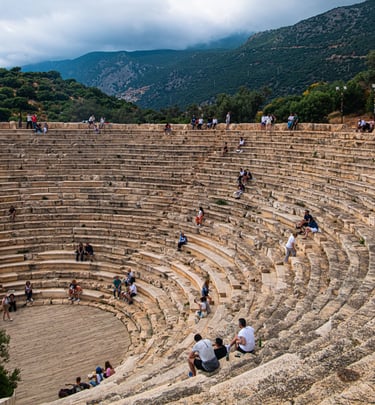 Tourists sitting on the stone steps of the ancient Greek theatre in Kas, Turkey, with mountain views.