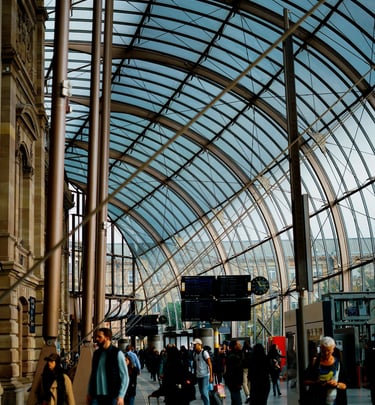 Commuters walk through a modern European train station under a large glass vaulted ceiling.