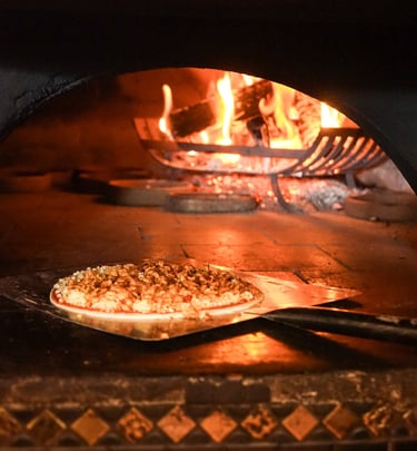 Authentic wood-fired pizza baking on a metal peel inside a stone oven with roaring orange flames.