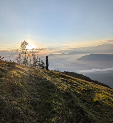 A hiker stands on a grassy mountain ridge watching the sunrise over a foggy valley.