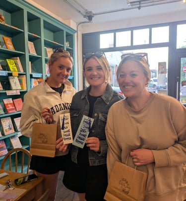 Three smiling women holding shopping bags and linocut bookmarks inside a cozy local bookstore.