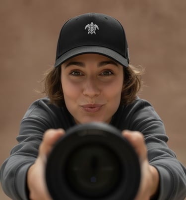 A smiling woman wearing a black sea turtle logo baseball hat holds a professional camera lens toward the viewer.
