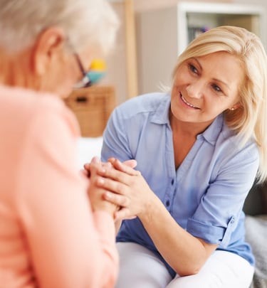 A blonde female caregiver provides compassionate home care while holding hands with an elderly woman.