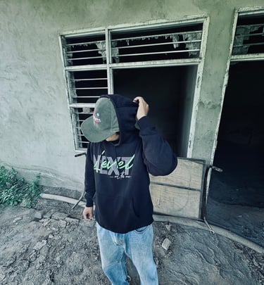 Man in black hoodie and corduroy cap standing outside a rustic, abandoned concrete building.