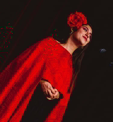 A female singer wearing a red flower in her hair and a traditional patterned red shawl performing on stage.