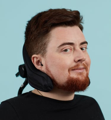 Jamie, a white person on a blue background, electric wheelchair headrest behind their head.