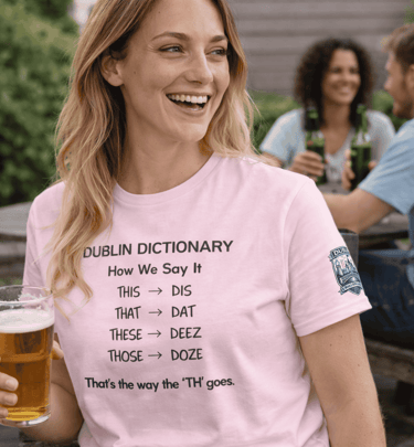 A smiling woman in a pink Dublin Dictionary t-shirt holding a beer at an outdoor patio.