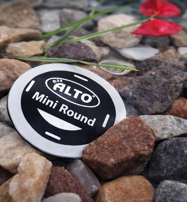 A waterproof Alto Mini Round security tag resting on smooth river stones in shallow water.