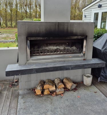 A modern outdoor concrete fireplace on a backyard deck with stacked firewood and a tall chimney.