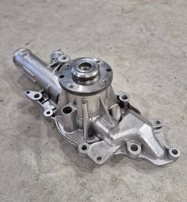 A new metallic automotive engine water pump replacement part sitting on a concrete workshop floor.