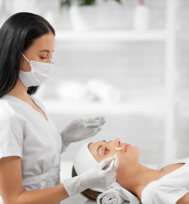 Professional esthetician in a mask applying a chemical peel facial treatment to a client in a spa.
