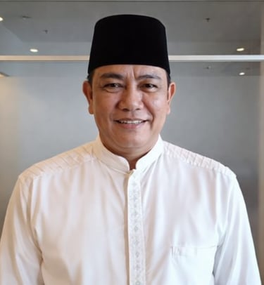 Smiling man wearing a traditional black songkok hat and white baju melayu shirt in an office setting.