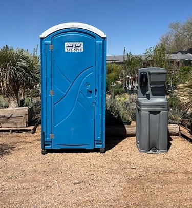 A blue portable toilet and a grey outdoor hand washing station on a gravel surface at a plant nursery.