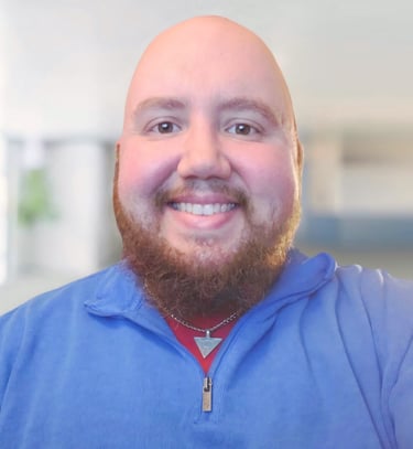 Smiling bald man with a red beard wearing a blue quarter-zip pullover and a silver necklace.