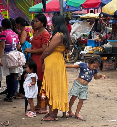 Indigenous women and children walking through a crowded outdoor marketplace with colorful stalls.