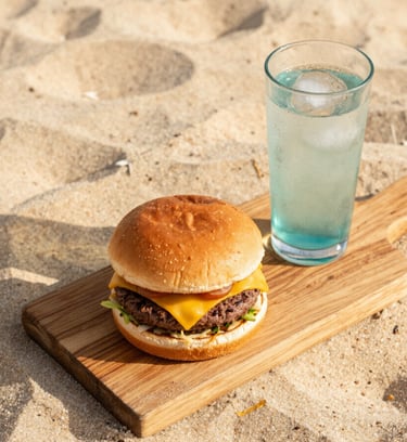 Overhead flatlay of a gourmet burger with melted cheese on a rustic wooden board, next to a glass of cold drink with condensation, soft golden beach light, high-end food styling