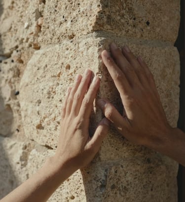 Close-up photography focusing on two people's hands gently touching a rustic limestone wall in a Southern European setting. Soft, warm sunlight creates a cinematic and authentic atmosphere with a shallow depth of field.