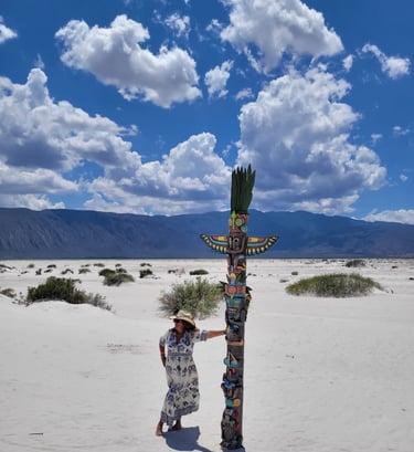 Una mujer con sombrero de sol posa junto a un colorido tótem en paisaje desértico de arena blanca.
