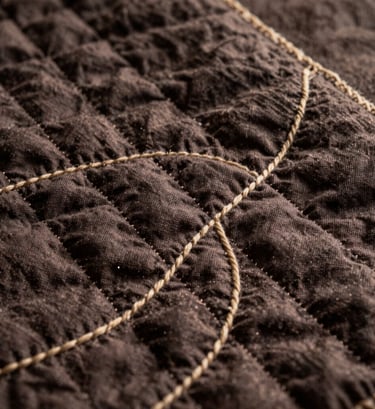 A macro photograph focusing on the meticulous hand-stitching of a quilt. The thread is a soft sand color, creating flowing, organic wave patterns across a deep espresso linen fabric. The lighting is warm and directional, highlighting the rich texture of the textile.