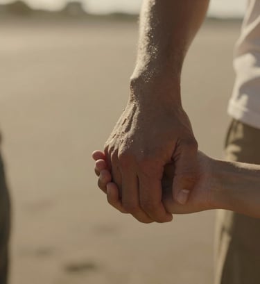Close-up cinematic shot of two people's hands gently intertwined, shot in a sun-lit outdoor setting in the North American / US. Warm, golden hour light, shallow depth of field, authentic lifestyle photography. Colors include soft sand and deep earth browns.