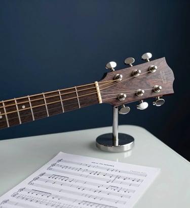 A close-up shot of an acoustic guitar neck and handwritten sheet music on a modern desk. The background is a solid deep midnight navy wall. The composition is clean and artistic, with cool steel grey metallic accents. A creative workspace in a Spanish city home.
