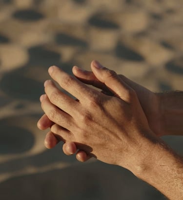 A candid close-up shot of two people's hands gently intertwined. The lighting is warm and golden, with soft sand highlights and deep charcoal shadows in the background. The mood is intimate and authentic.